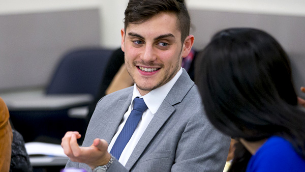 A male NYU GPH student in a gray suit and blue tie is smiling and gesturing while talking to a female student in a blue outfit.