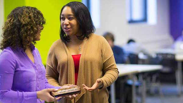 Two in-person NYU Master’s in Public Health students are standing together and conversing in a study area.