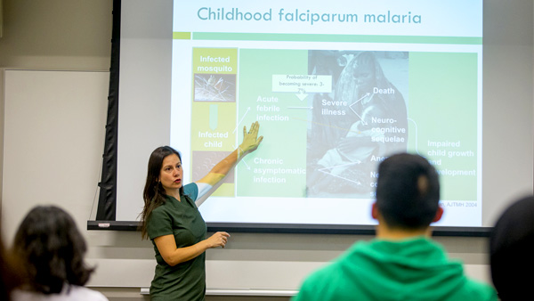 NYU GPH Associate Professor of Global and Environmental Health, Yesim Tozan, is standing at the front of a classroom while lecturing to in-person MPH students and placing her hand on a projector screen showing a slide about childhood falciparum malaria.