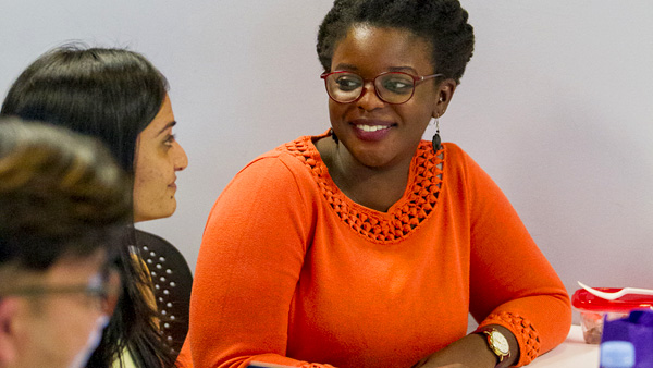 Two in-person NYU MPH students are sitting together in a classroom and looking at each other.