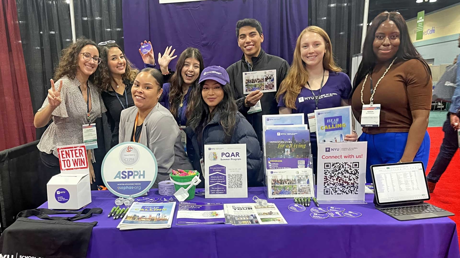 A diverse group of eight Master of Public Health (MPH) students are smiling and posing behind a table at an NYU GPH admissions event.