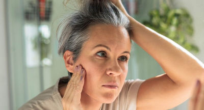 woman looks at greying hair and skin in mirror