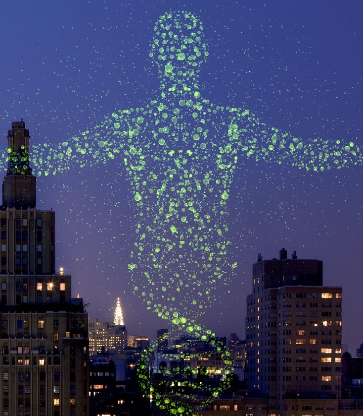 A glowing green silhouette of a person with outstretched arms, composed of floating dots, stands above New York City buildings at night.